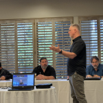 Man standing and speaking to a seated group in a conference room with laptops on a table and wooden blinds in the background