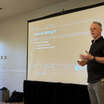 Speaker in a black shirt stands beside a projector screen showing a slide titled 'Directly Selling AI' with bullet points and hexagonal graphics in a conference room.