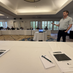 Presenter at the front of a conference room giving a slide presentation while attendees sit around a long, U-shaped table behind white tablecloths.