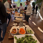 People line up at an outdoor buffet under beige canopies, serving salads and bread from a long wooden table.