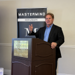 Speaker in a dark suit gives a presentation at a podium, with a Mastermind Peer Groups banner behind him.