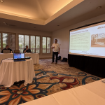 Speaker in a white polo presents beside a projected slide in a conference room with attendees seated at a long table; slide shows 'The Results Software journey' and a building image, plus a 'Results' logo in the corner.