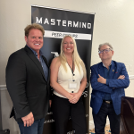 Three smiling adults stand in front of a vertical banner reading 'MASTERMIND' with 'PEER GROUPS' below, posing for a photo.