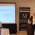 Female presenter in a blazer speaks beside a projected slide in a conference room, with a tall 'Mastermind Peer Groups' banner nearby.